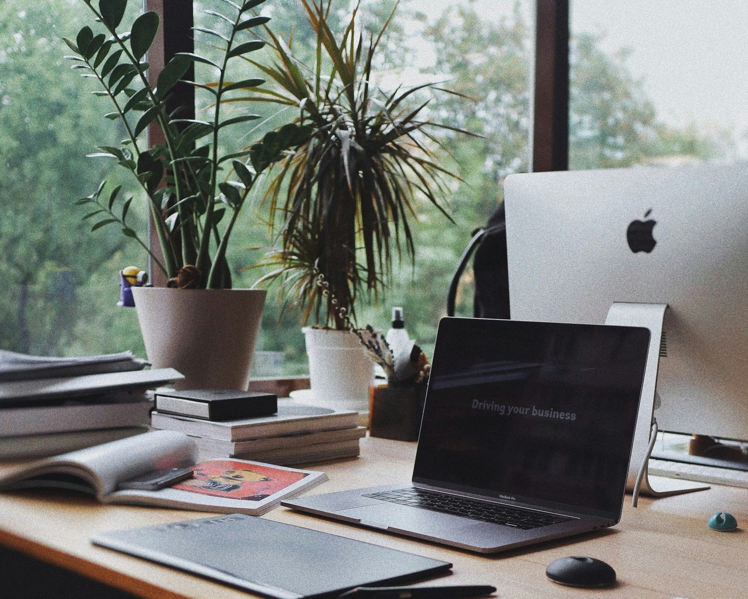 A laptop on a wooden desk by a window with plants and notebooks